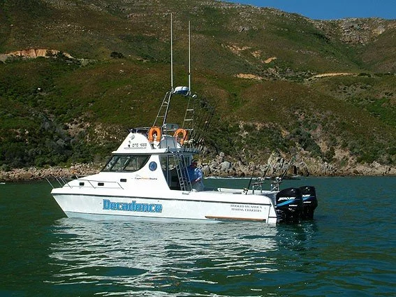 A white boat named 'Decadence' with two large outboard motors, floating on calm water with a hillside in the background.