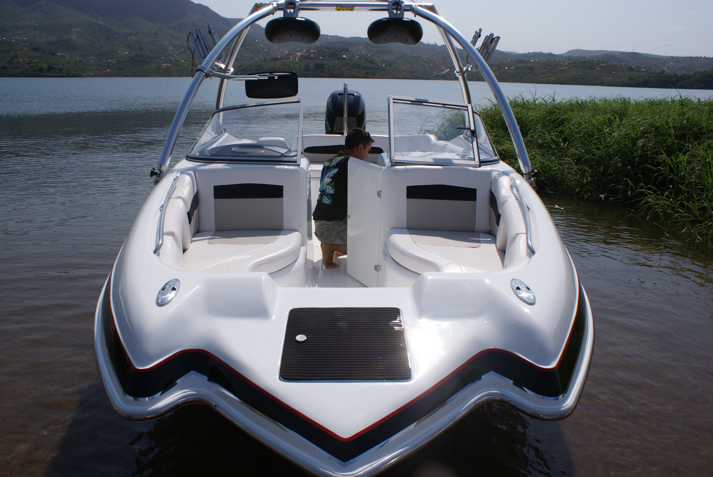 A man in shorts and a dark T-shirt is kneeling on the deck of a white boat with cushioned seating, looking into the cabin area, with water and grassy shoreline on either side and hills in the background.