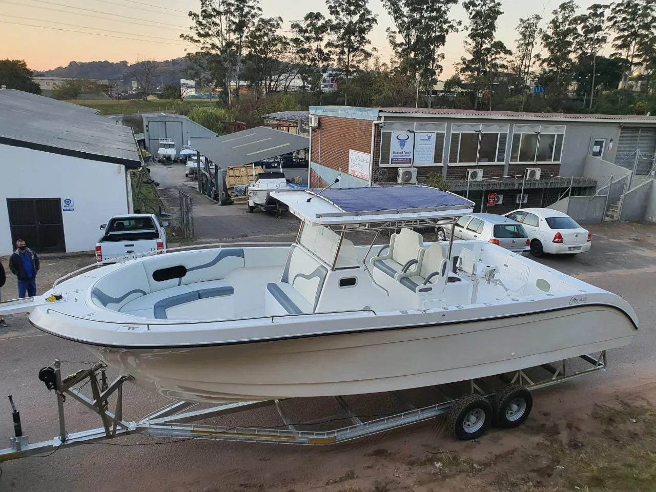 A white motorboat with a blue canopy on a trailer in a parking lot, with buildings and trees in the background.