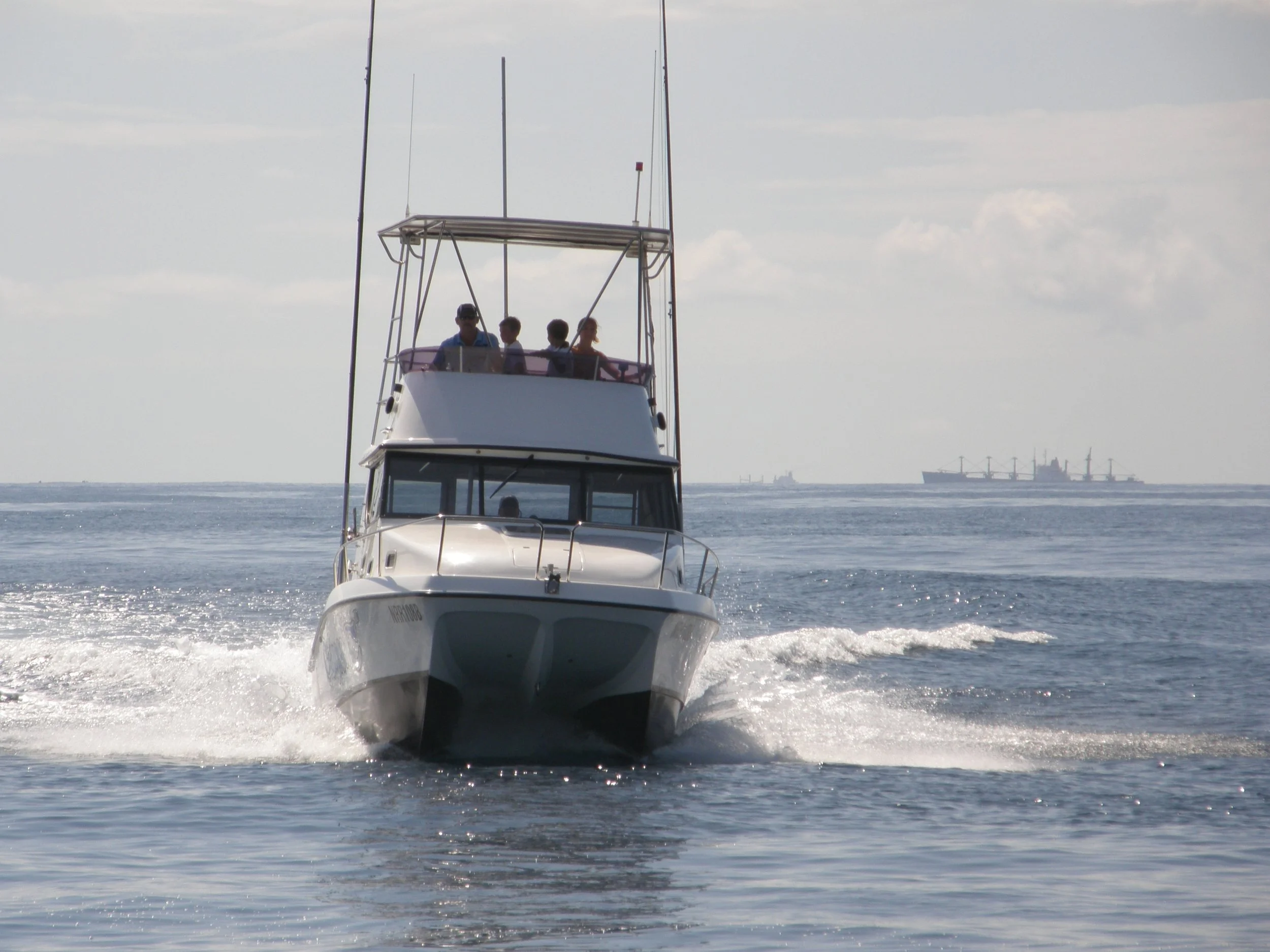 A white motorboat moving fast on the water with four people on the upper deck and two people visible through the front window, with a large cargo ship and a cruise ship in the background on a partly cloudy day.