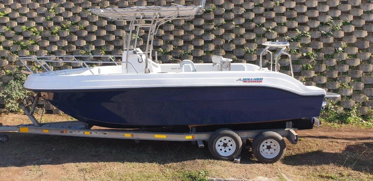 A white and blue boat on a trailer on a dirt ground in front of a stone retaining wall with plants.