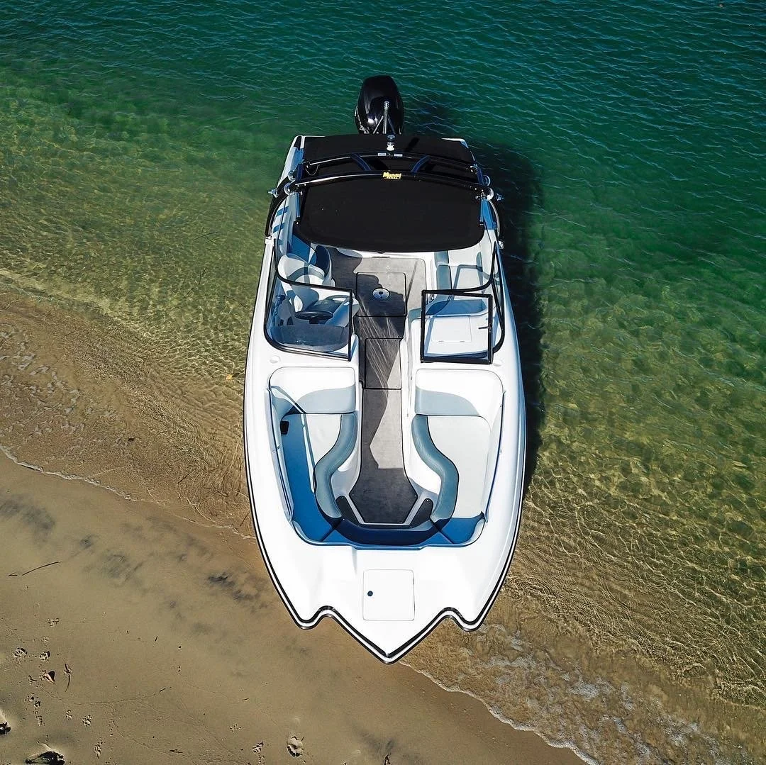 An aerial view of a white speedboat docked on a sandy beach, with clear green water surrounding it.
