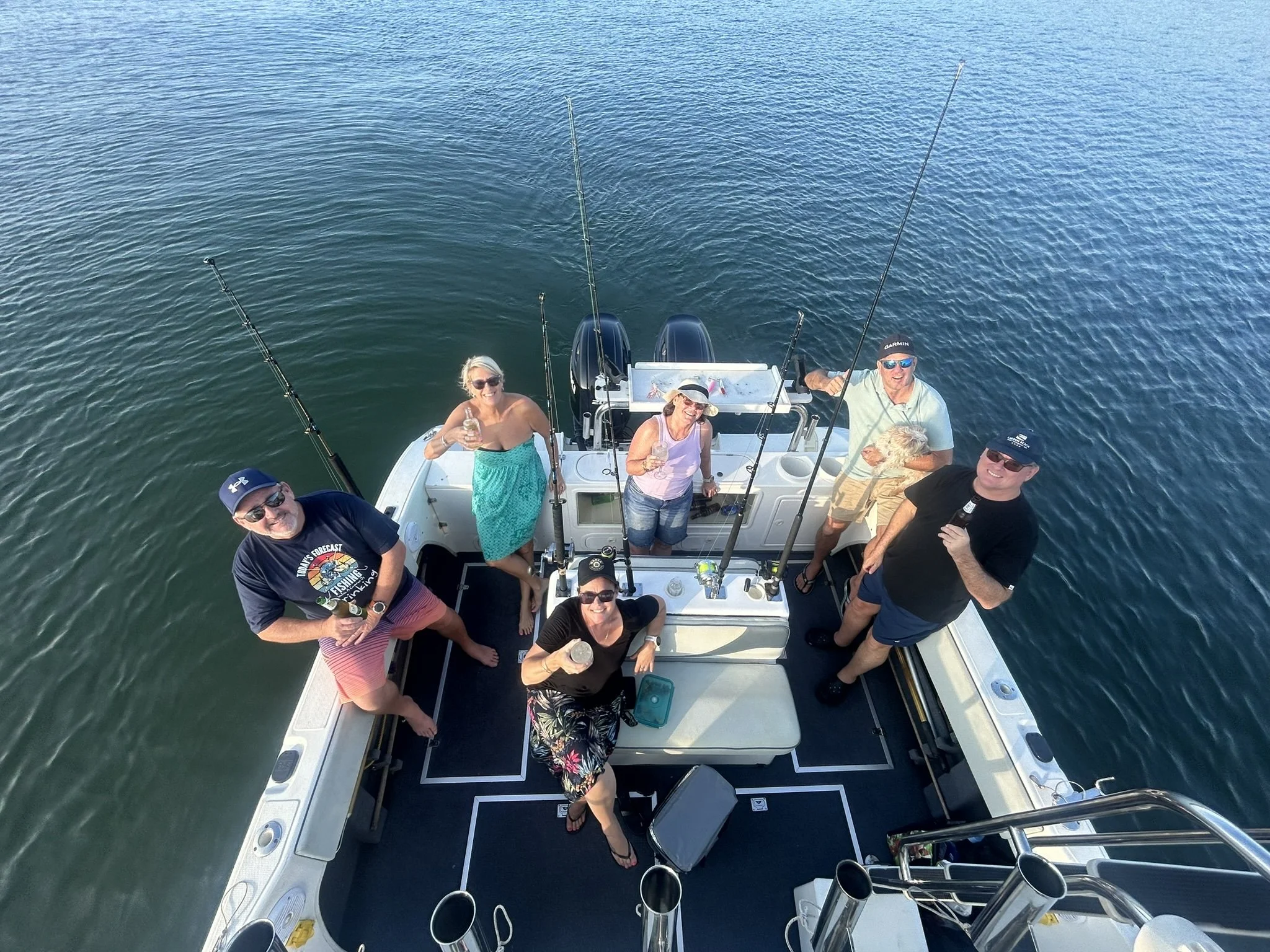 Six people on a boat enjoying a sunny day, with some holding drinks and fishing rods, on calm water.
