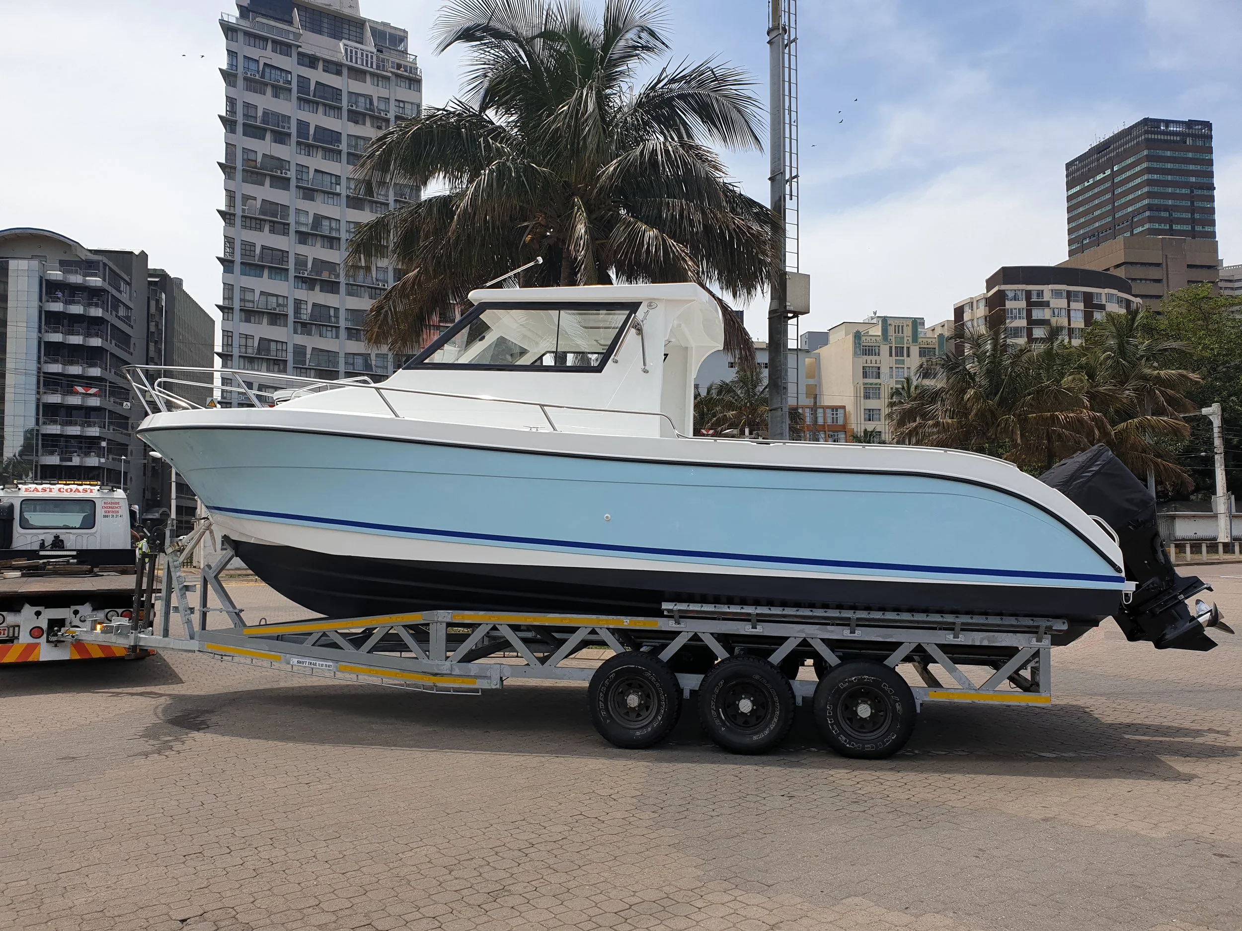 A white and blue motorboat on a trailer in an urban harbor setting with high-rise buildings and palm trees in the background.