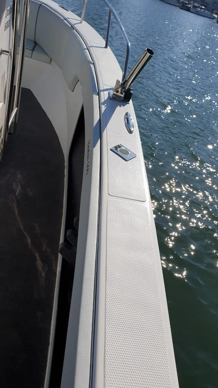 A close-up view of the side of a white boat with a metal railing, a metal cleat, and a cylindrical metal pipe, on calm water with sunlight reflecting on the surface.