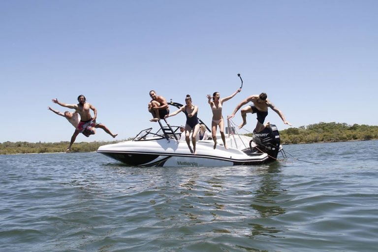 Group of five people on a boat in a body of water, with one person mid-air jumping off the boat, enjoying a day of water recreation.