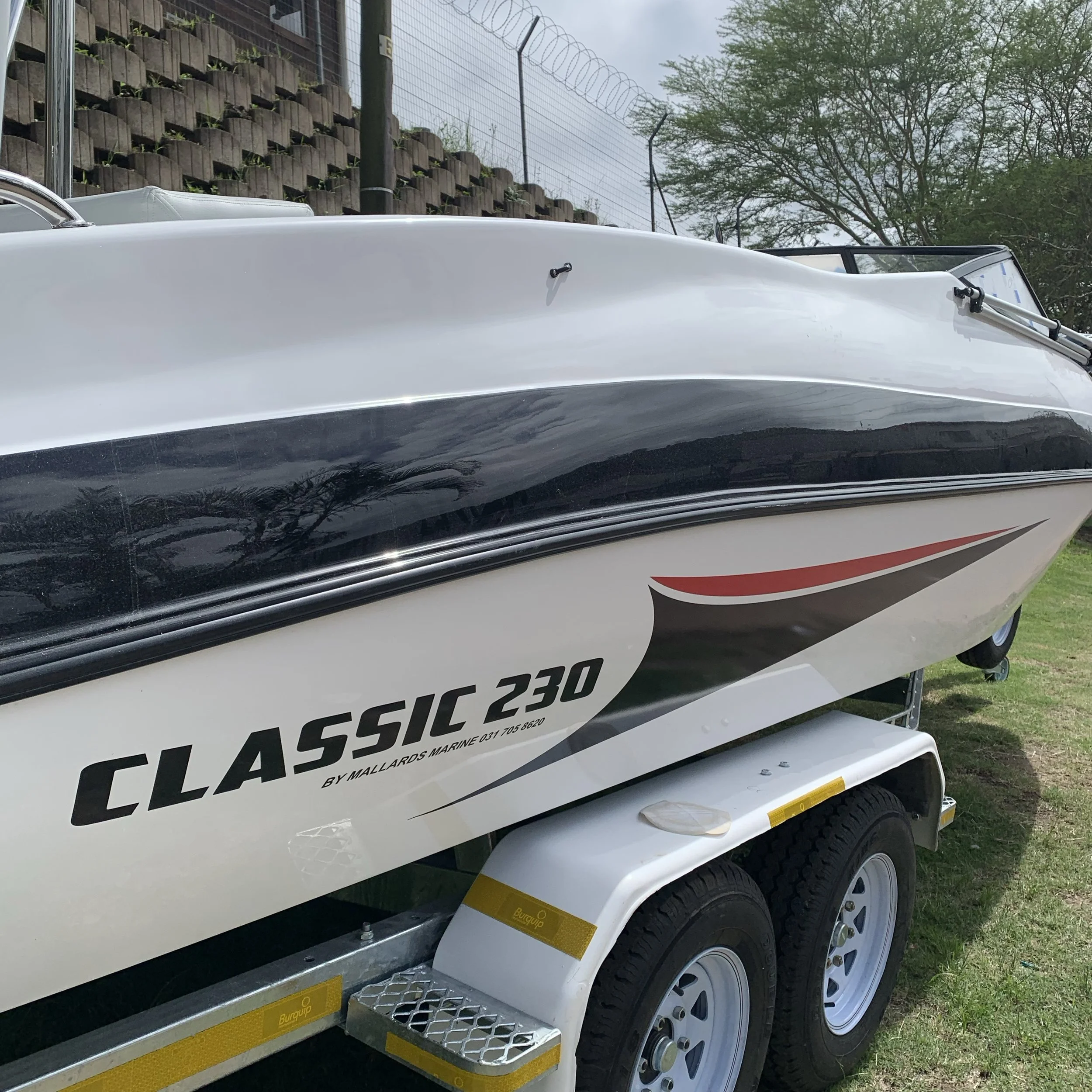 A white and black speedboat named Classic 230 on a trailer with yellow reflective markings parked on grass, with trees and a fence in the background.