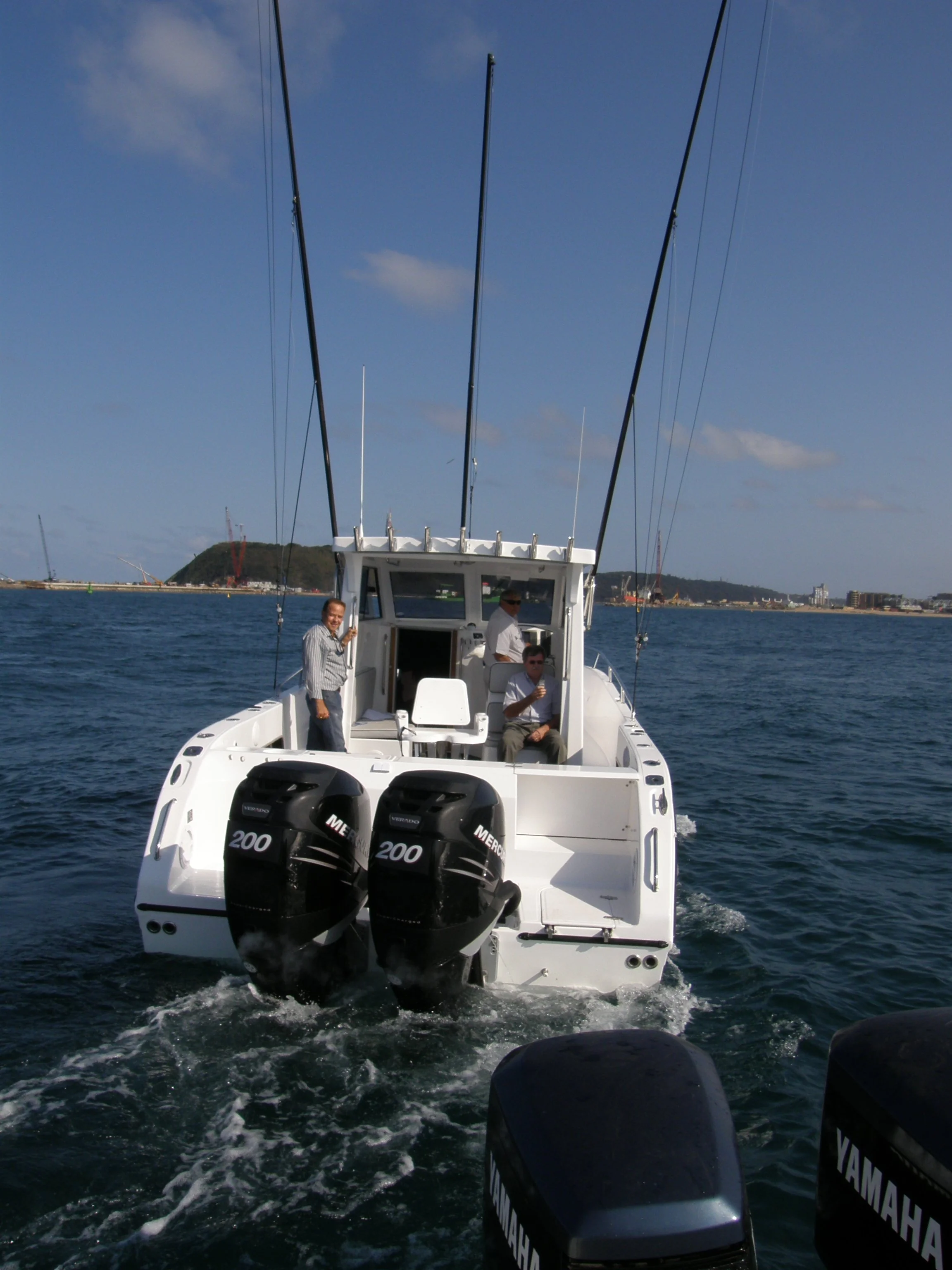 A white motorboat on the water with three people on board, two men standing and one seated, with two black outboard engines at the stern, and a coastline with buildings and a hill in the background under a blue sky.