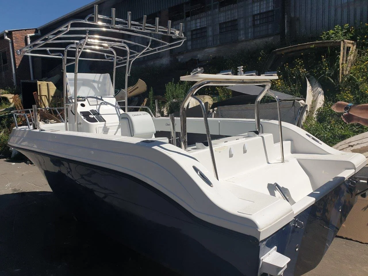 White and blue center console boat with a T-top, accessories and seating, parked on land near plants and a wooden structure.