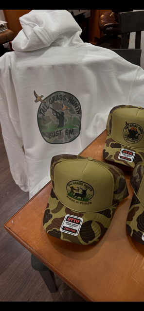 A white hoodie with a circular patch displaying a duck and the words 'TALL GRASS HUNTER BUST EM'. In front of the hoodie, there are camouflage baseball caps with a similar patch, all placed on a wooden table.