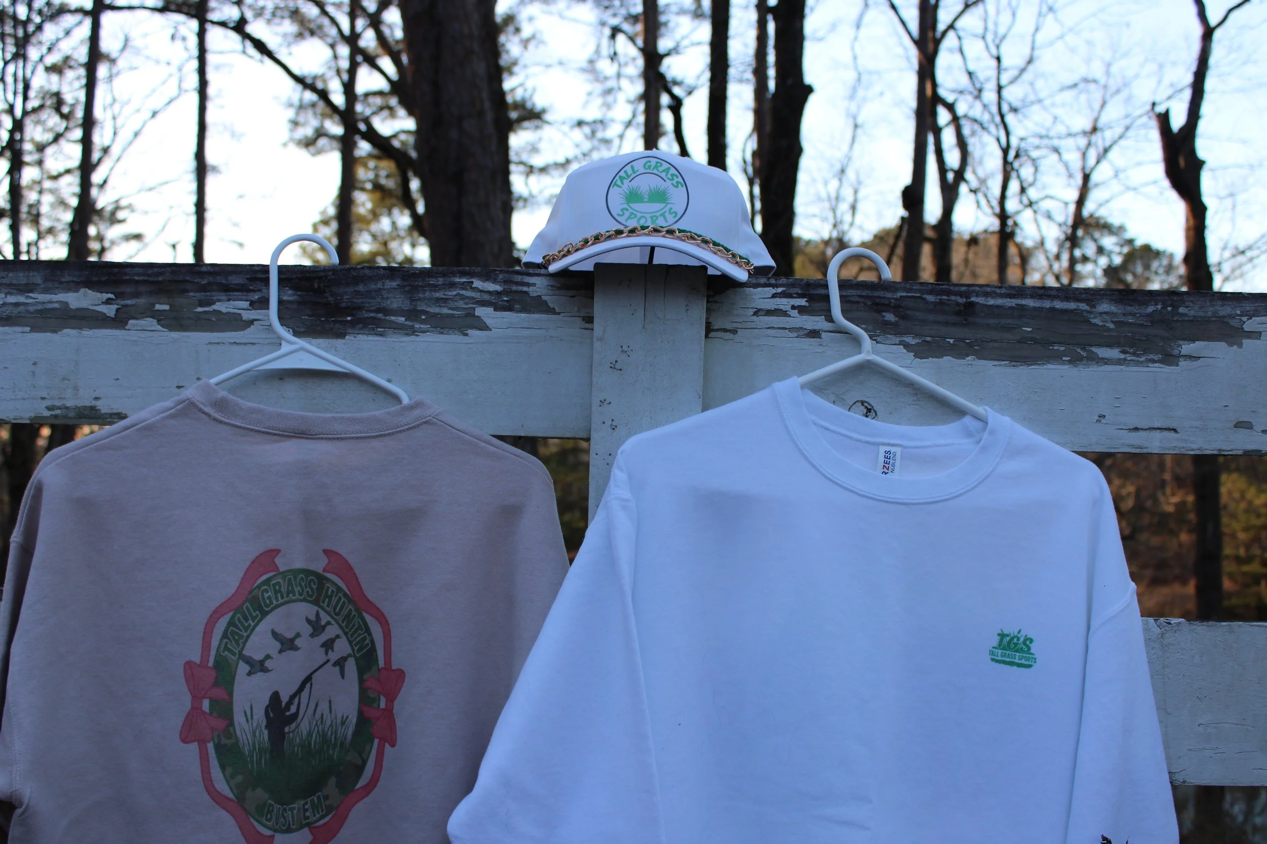 A white baseball cap with green and blue embroidery that reads 'Tall Grass Sports' is placed on a wooden beam outdoors with trees in the background. Two white T-shirts hang on the beam below the cap.