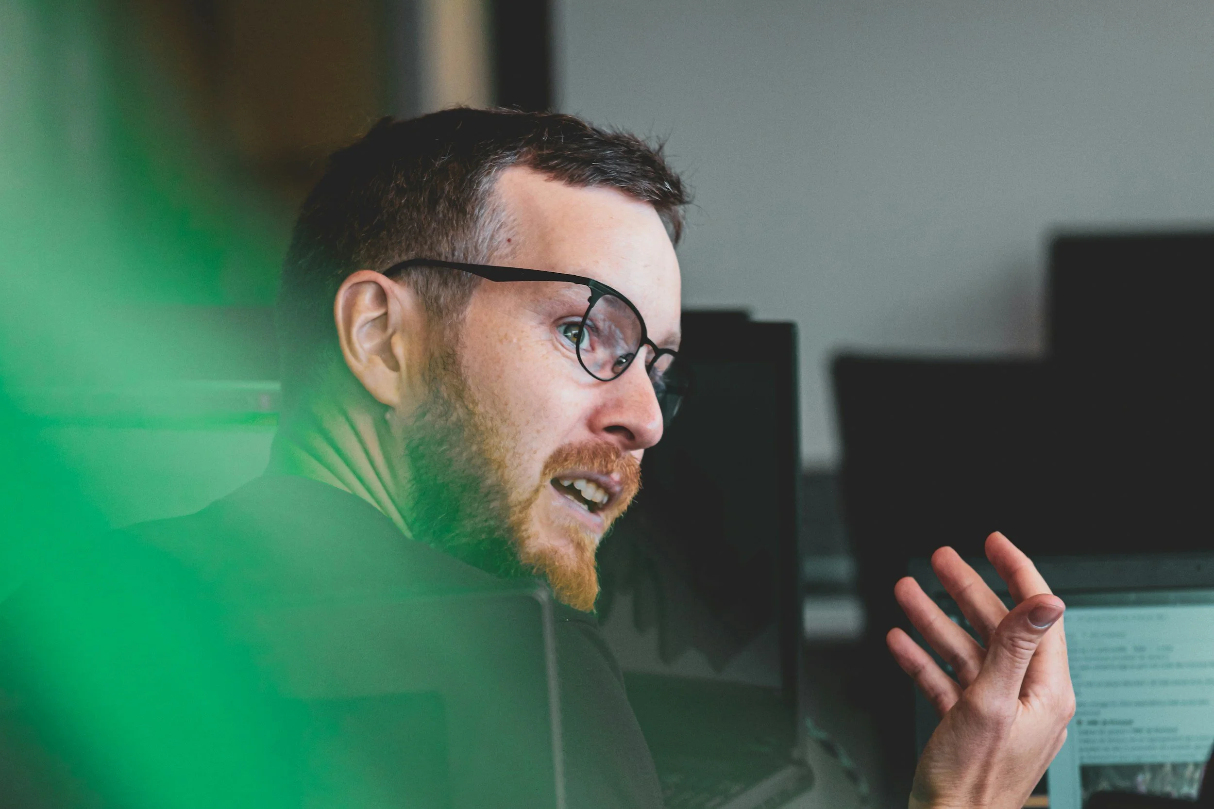 A man with glasses, a beard, and short hair speaking while sitting in front of computer monitors in an office.