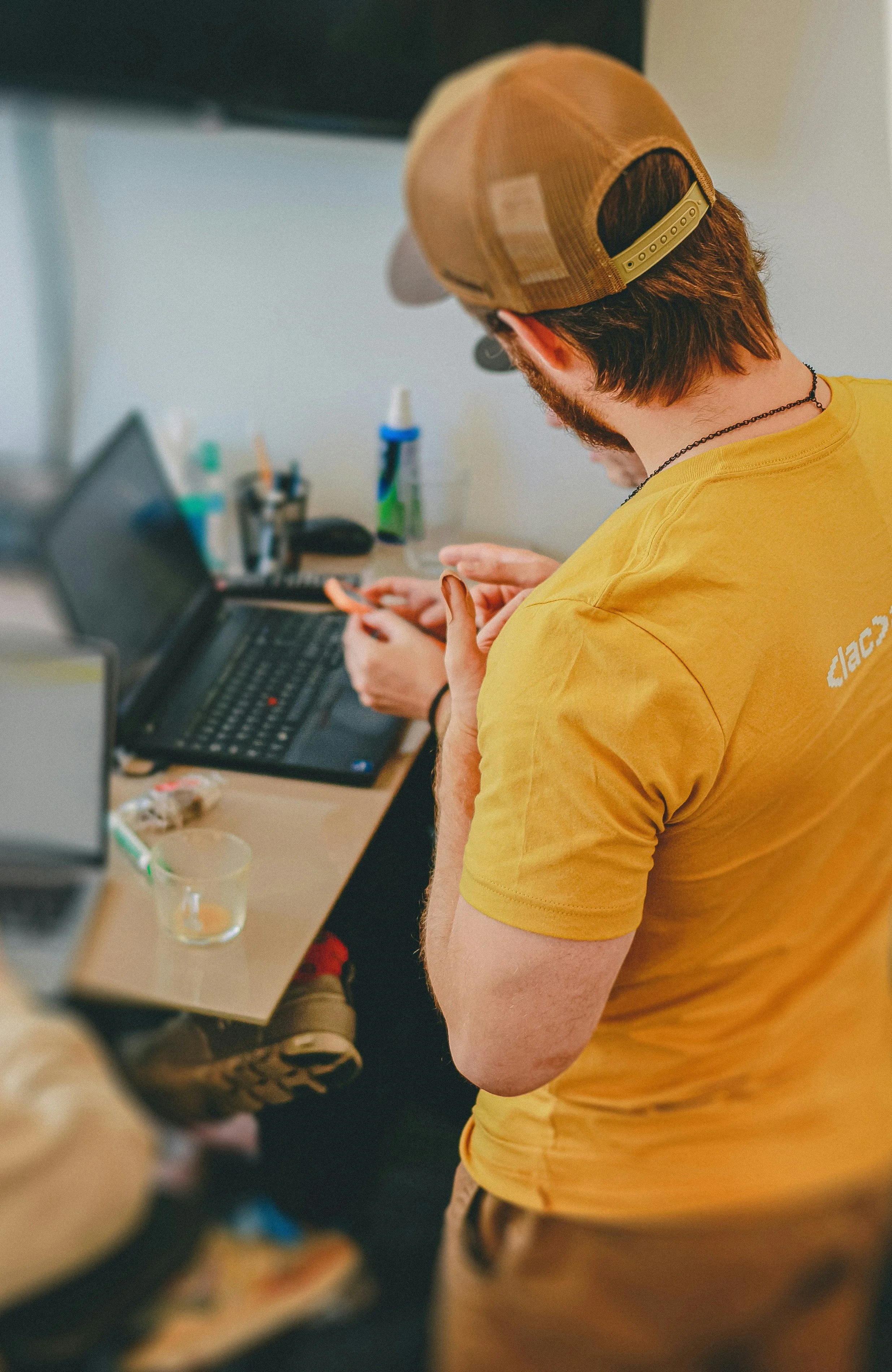 A man wearing a yellow T-shirt and a tan baseball cap is looking at his smartphone while working at a desk with a laptop, bottles, and other items.