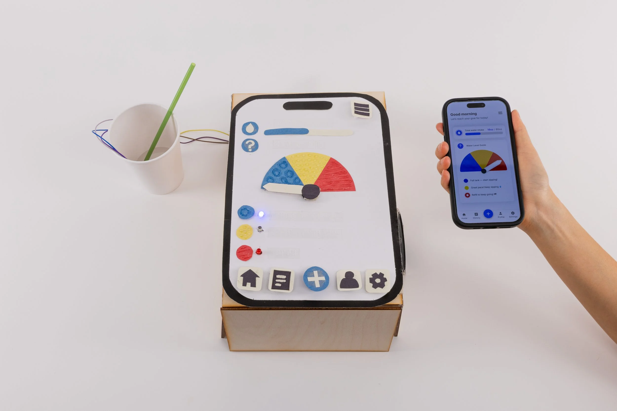 A tactile hydration-tracking prototype sits beside a white cup with a green straw connected by wires. The dashboard features Braille labels, textured color sections, and a lit blue LED. A hand holds a smartphone showing the Sip n’ Sense app with a ma