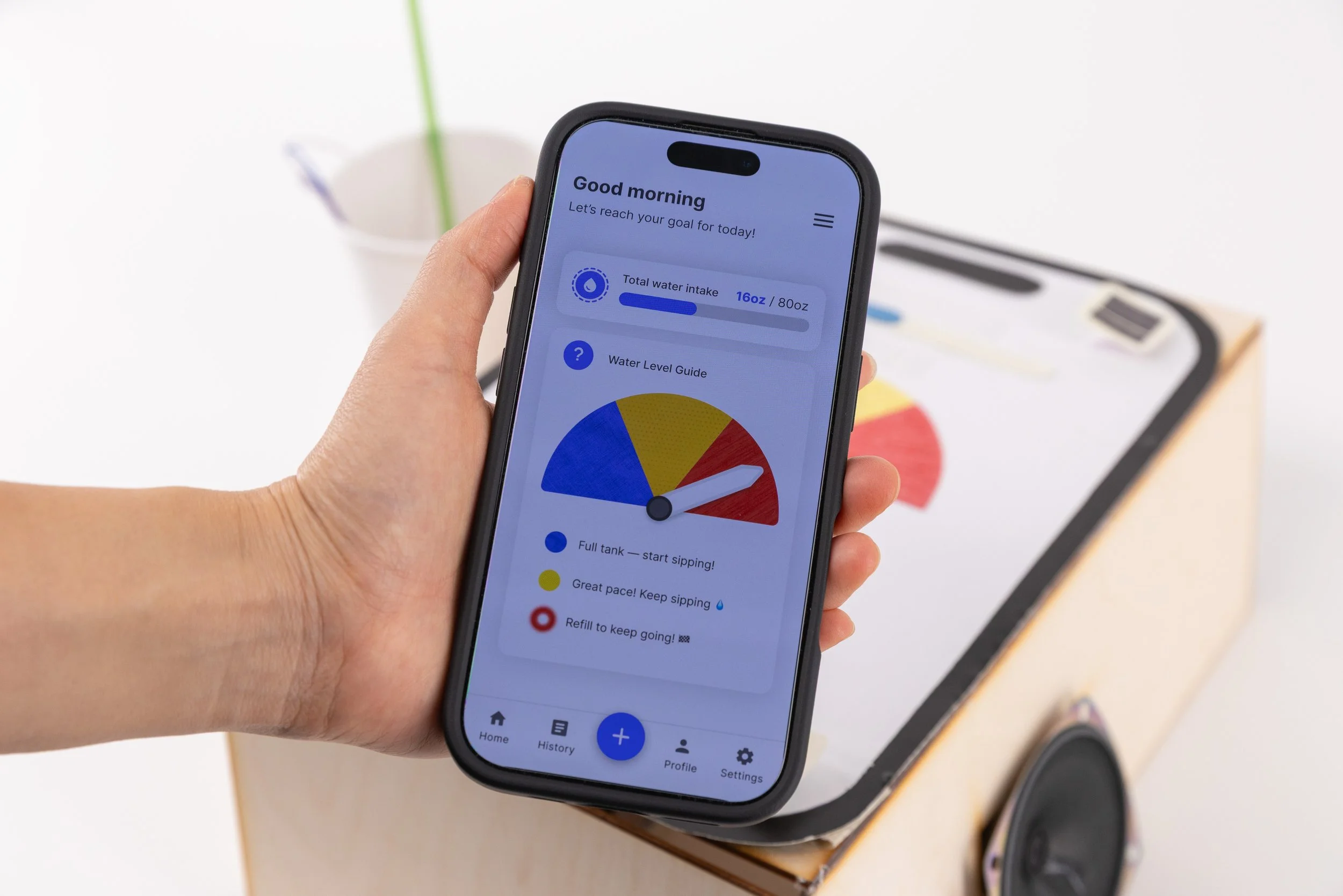 A person holds a smartphone showing the Sip n’ Sense app interface with a semicircular gauge and color-coded hydration levels. The physical prototype with matching tactile indicators is visible in the background.