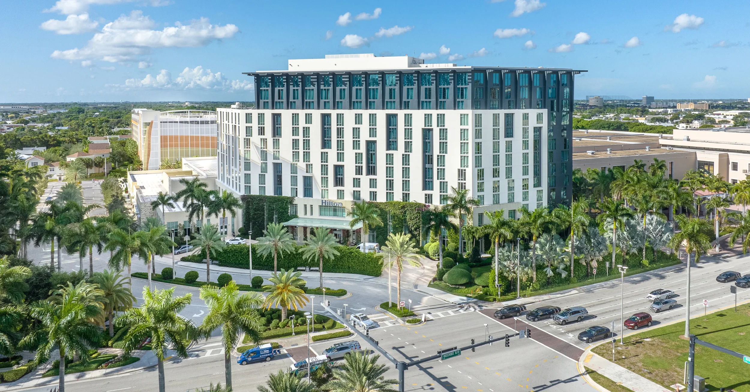 A tall Hilton hotel surrounded by palm trees, a busy street with cars, and green landscaping under a partly cloudy blue sky.