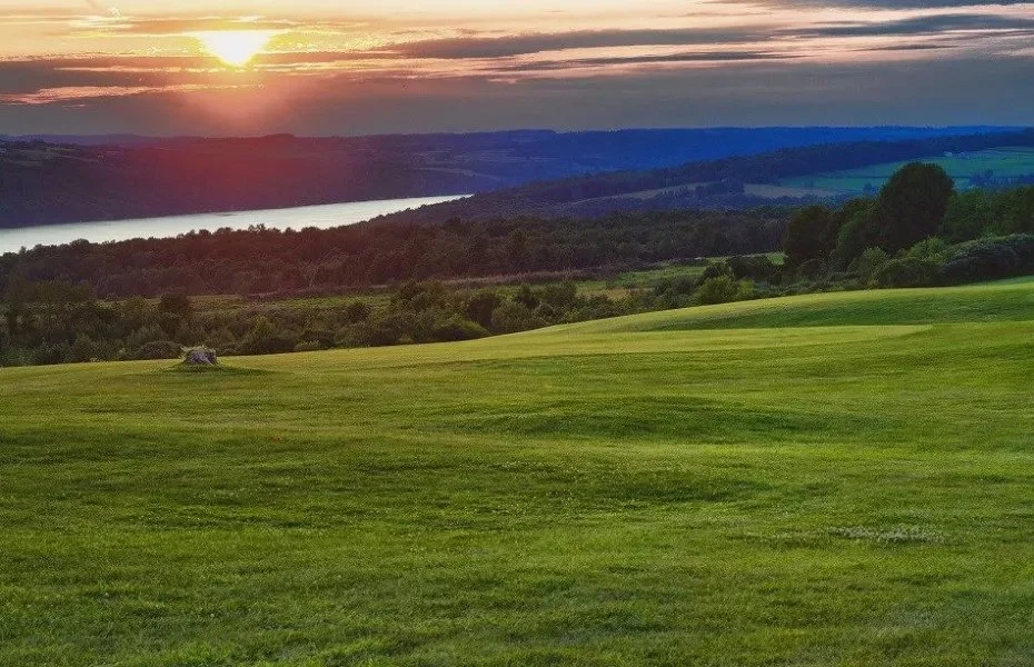 Sunset over lush green golf course with rolling hills and a body of water in the background.