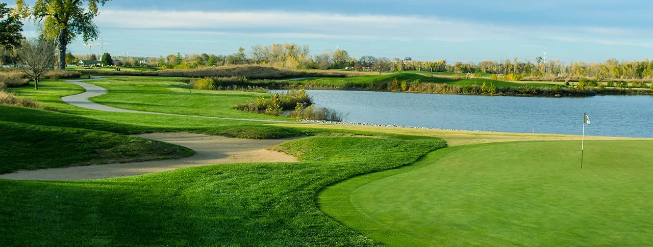 A scenic golf course with a putting green, sand bunkers, a water hazard, and a winding path through lush green grass, under a partly cloudy sky.