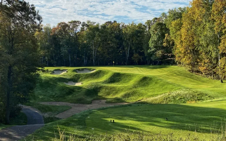 A golf course with a putting green, sand bunkers, and a surrounding wooded area under a partly cloudy sky.