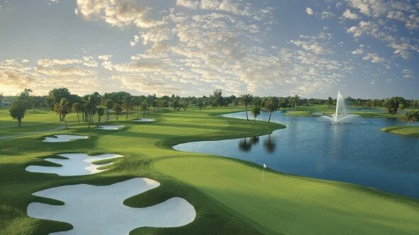 A scenic golf course with green fairways, multiple sand traps, a large water hazard, and a fountain in a pond, under a partly cloudy sky.