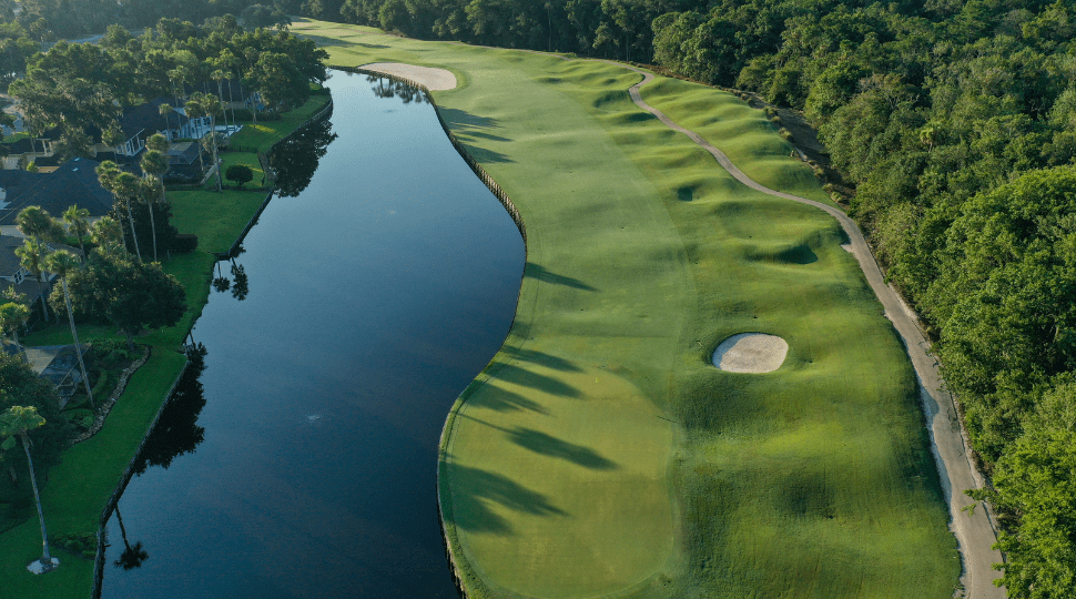 Aerial view of a golf course with a water hazard on the left and lush green fairways on the right, surrounded by trees.