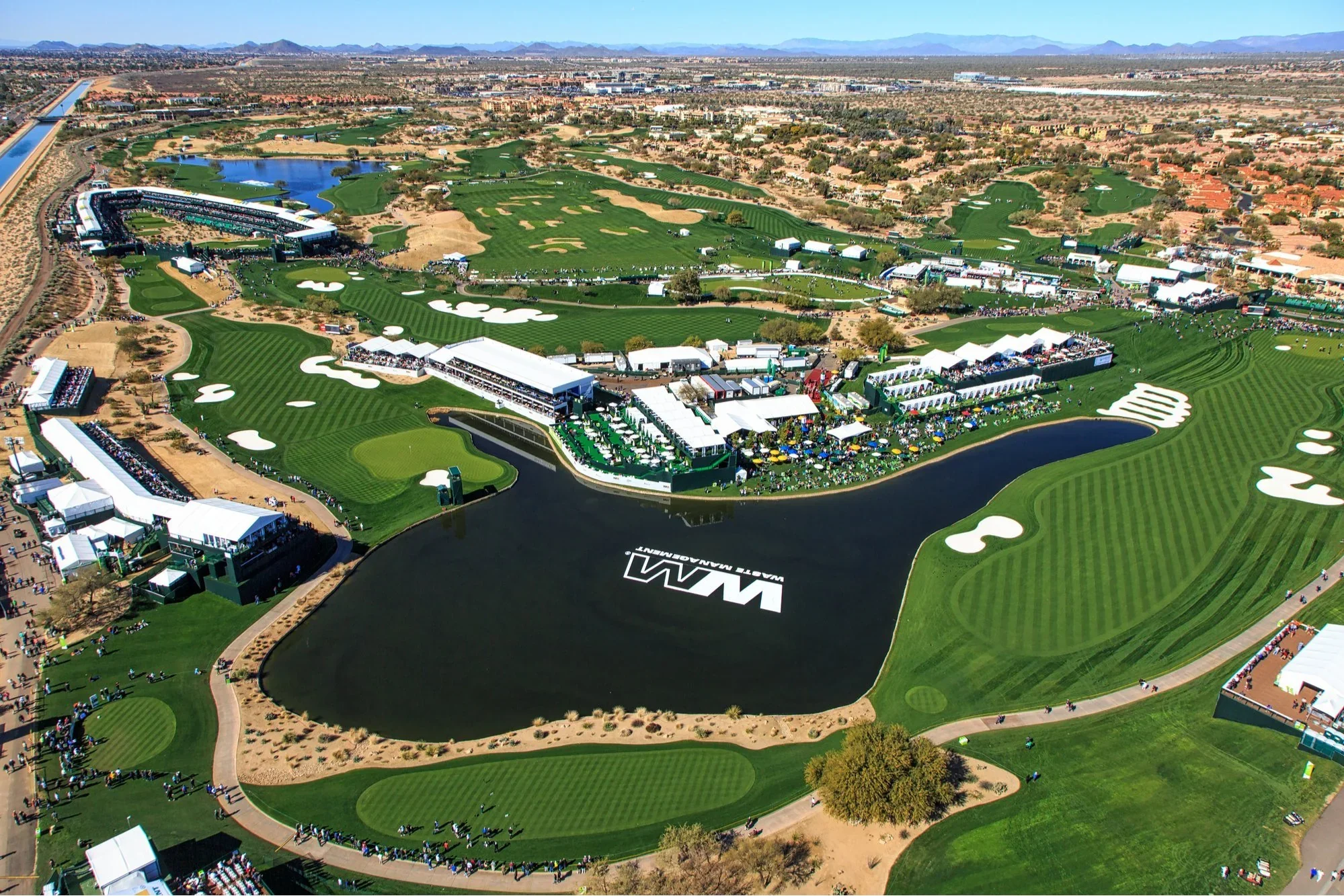 Aerial view of a golf tournament showing a golf course, water hazards, tents, and crowds of spectators at the Waste Management Phoenix Open.