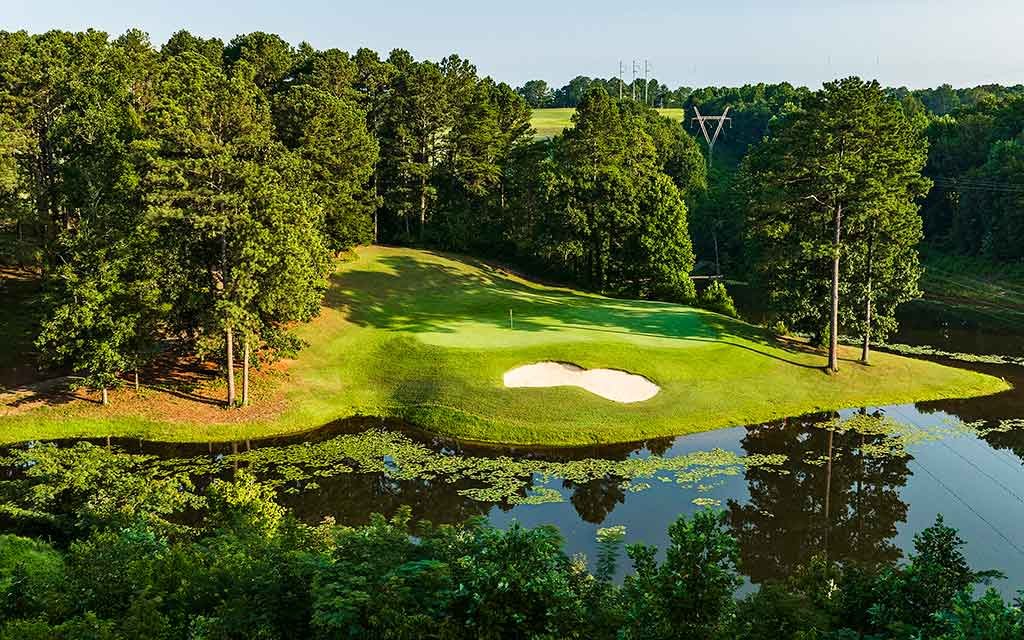 A scenic golf course fairway with a sand trap near a water hazard, surrounded by trees and lush greenery, under a clear sky.