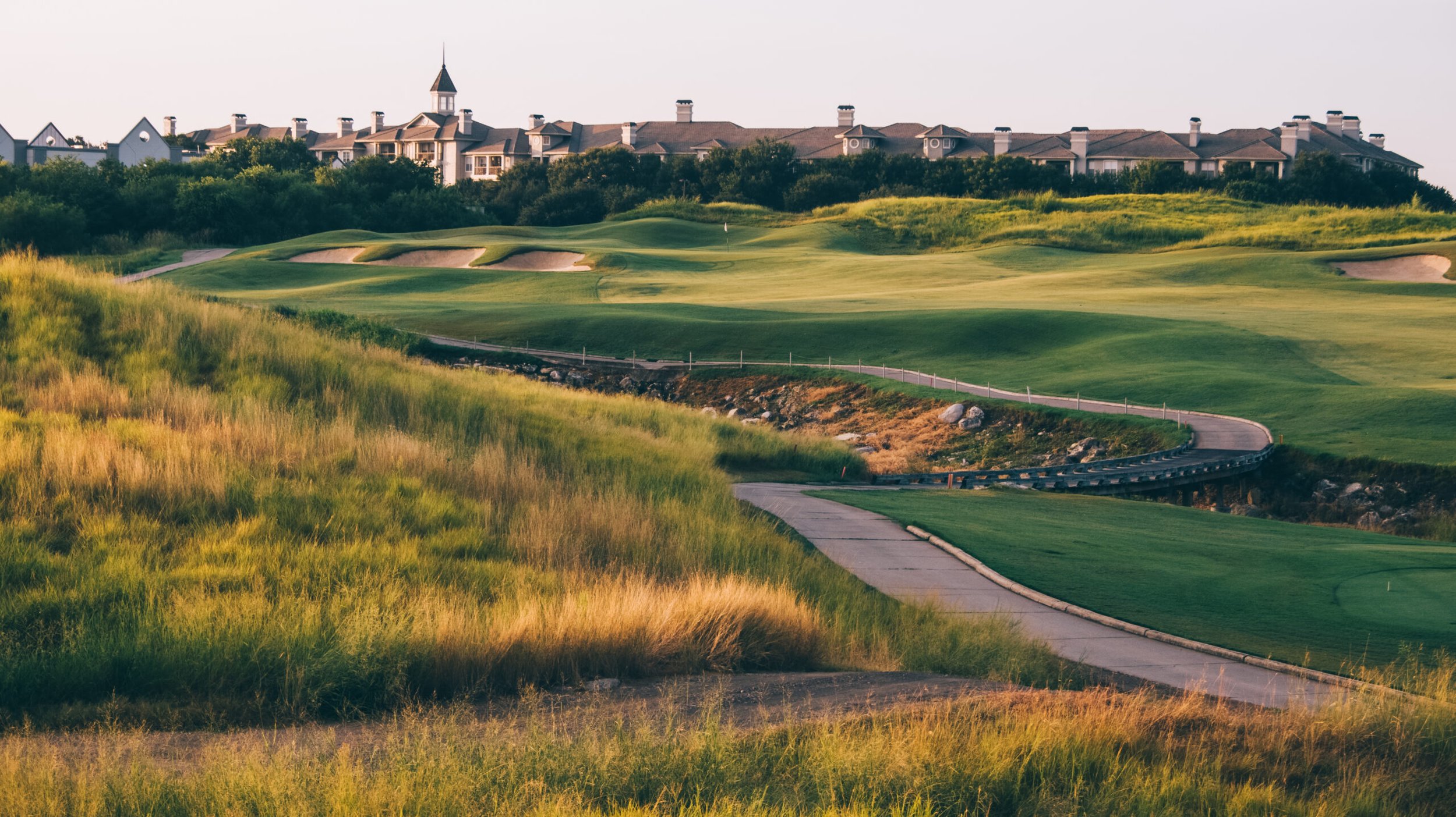 A scenic view of a golf course with rolling green hills, sand traps, a paved pathway, and houses with a turret in the background.