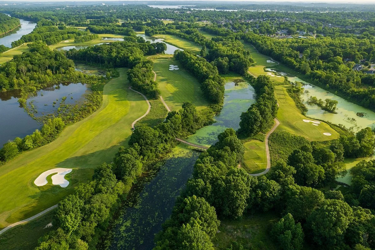 Aerial view of a lush green golf course with multiple water hazards, sand traps, and tree-lined fairways, set in a heavily wooded area.