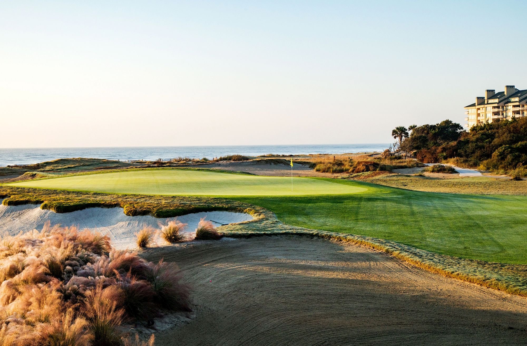 A scenic golf course near the ocean with sand traps, green fairways, and a clubhouse or hotel in the background under a clear sky.
