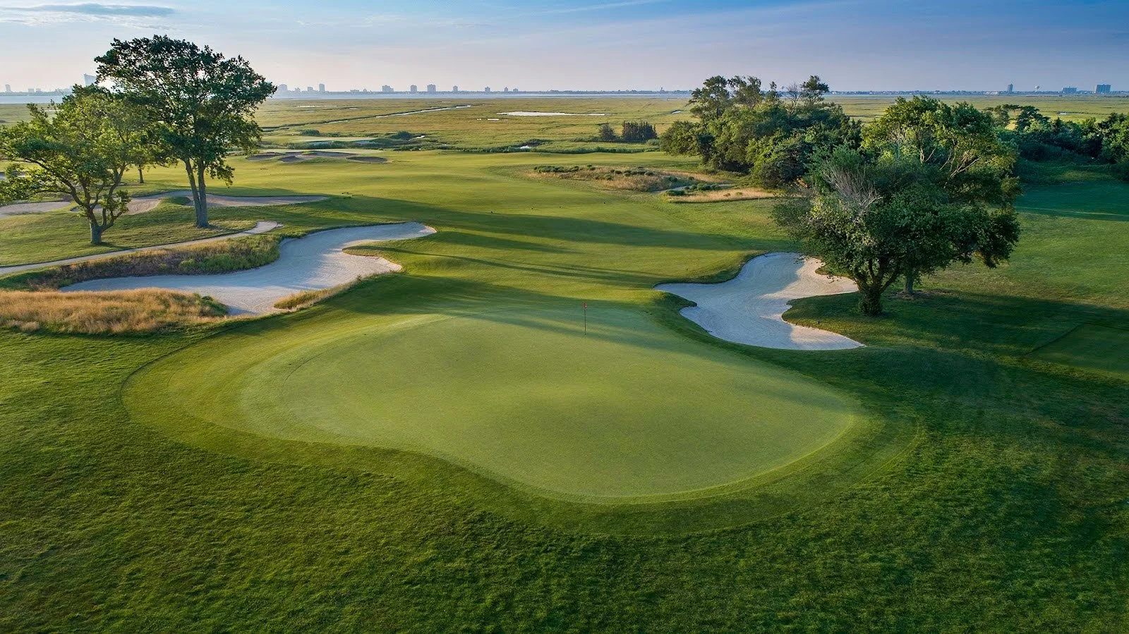 Aerial view of a golf course with green fairways, sand bunkers, and trees, with city skyline in the distance.