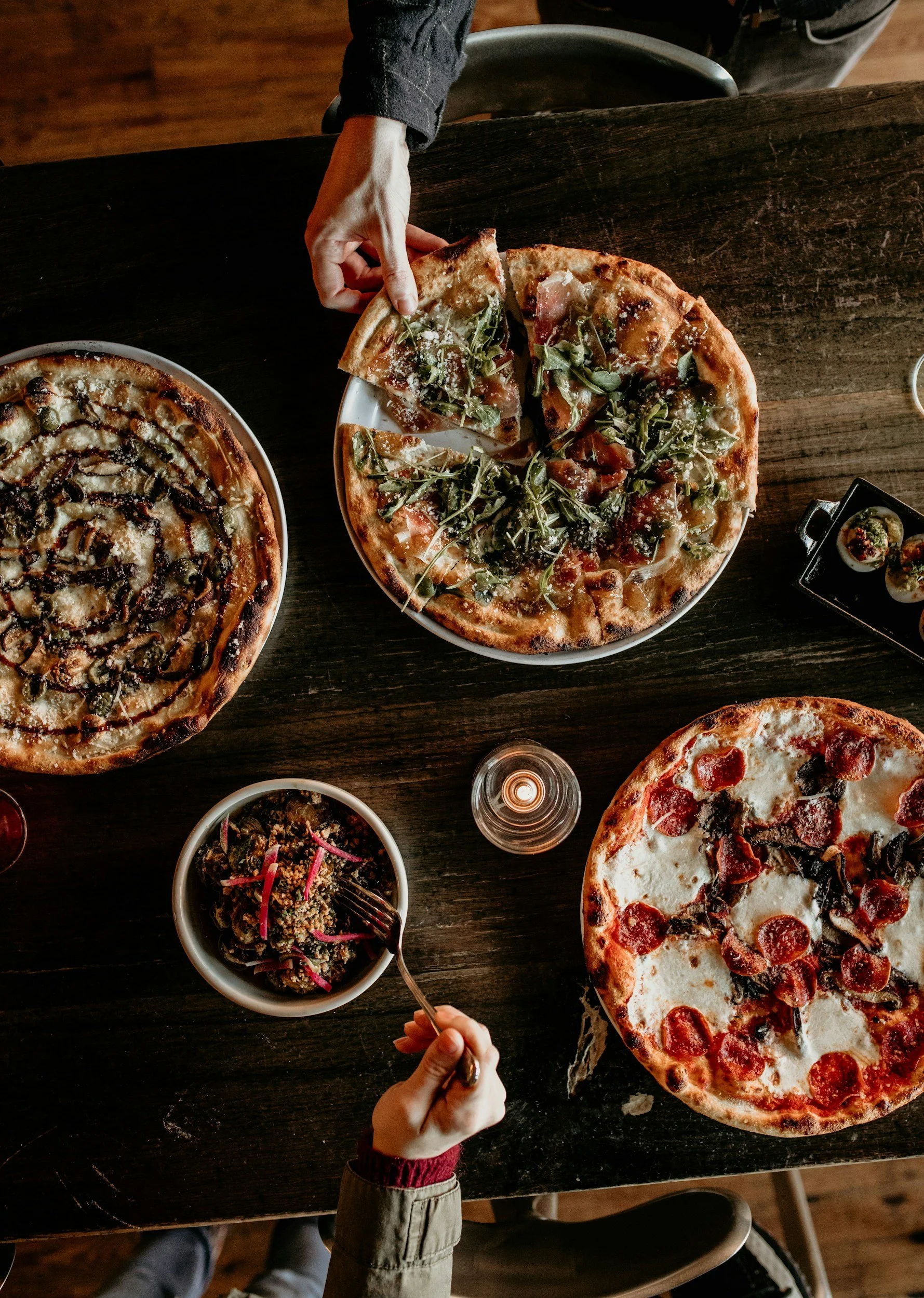 Top-down view of a dark wooden table with three pizzas, a bowl of pasta salad, a water glass, and two people reaching for slices at a cozy restaurant.