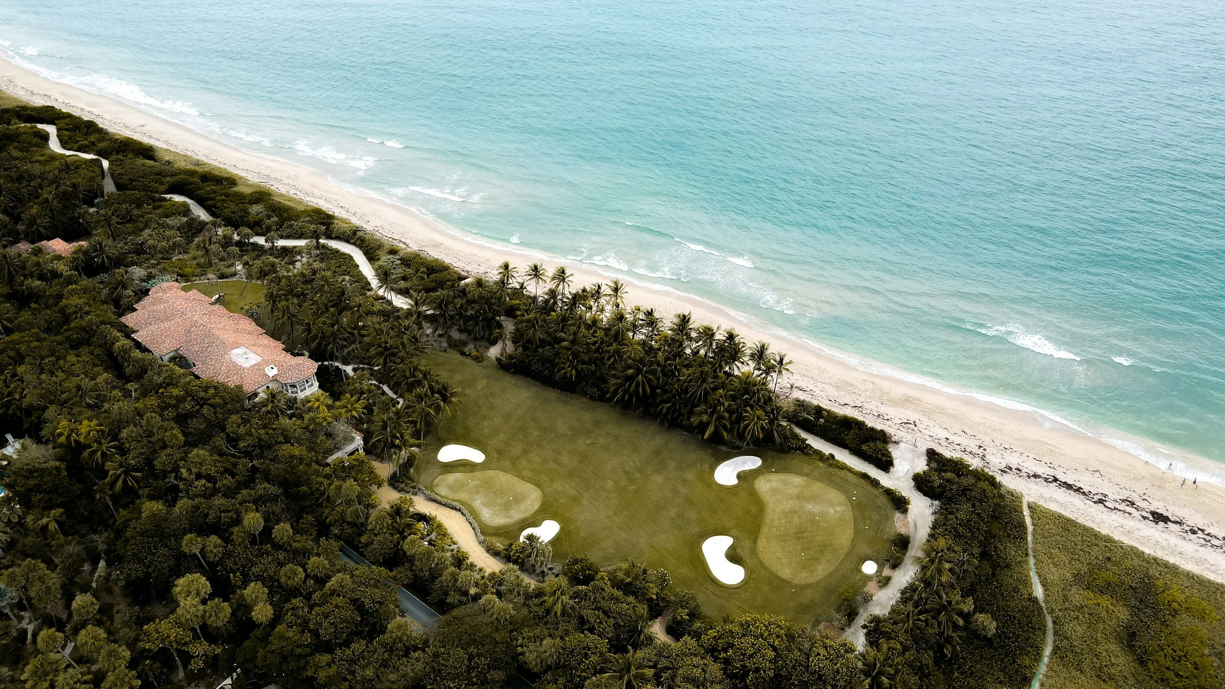 Aerial view of a coastal golf course with sand traps, green fairways, bordered by dense trees, next to a beach with white sand and light blue ocean waters.