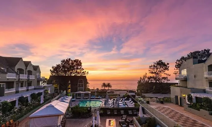 Sunset over a resort pool area with buildings, palm trees, and the ocean in the background.