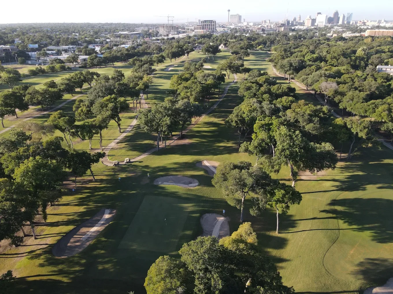An aerial view of a golf course with green fairways, sand traps, and trees, with a city skyline in the background.