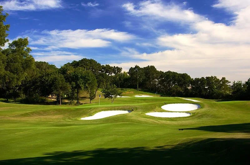 A golf course with sand bunkers, green grass, trees, and a blue sky with clouds.