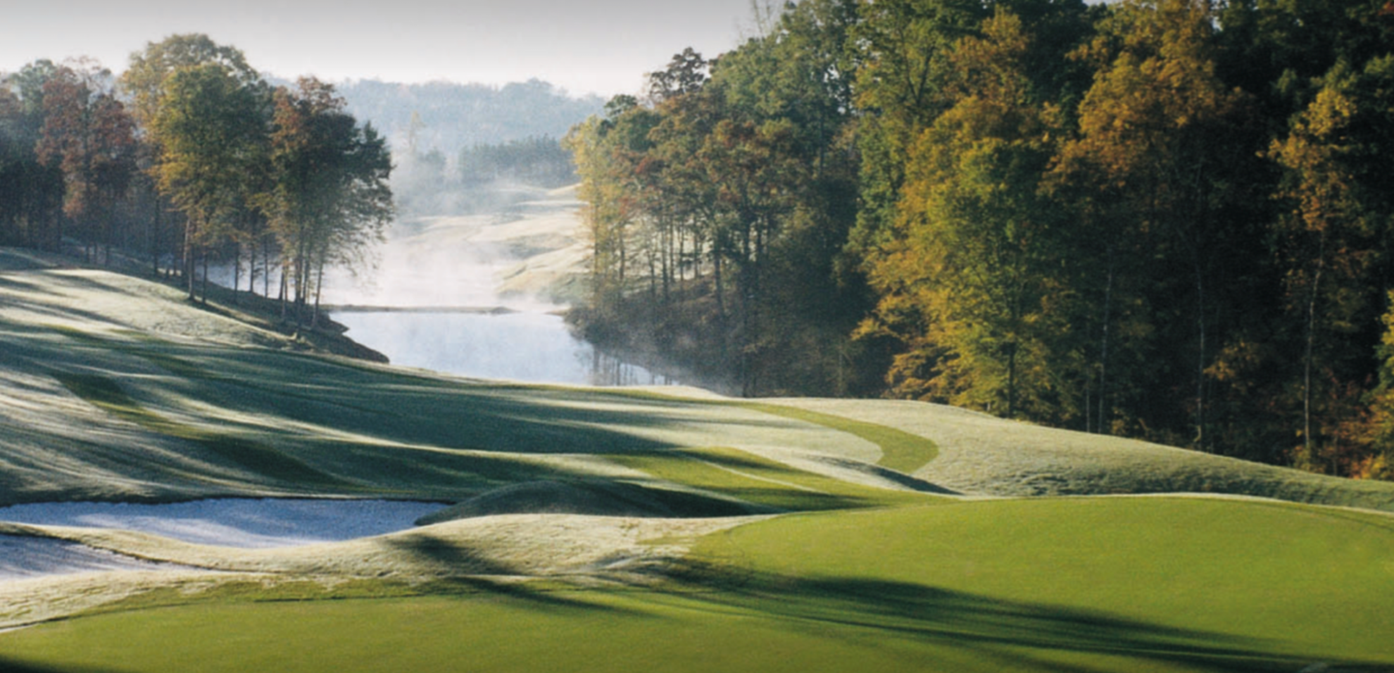 A scenic view of a golf course with rolling green fairways, surrounded by trees with fall foliage, and a river or lake in the background under a misty sky.
