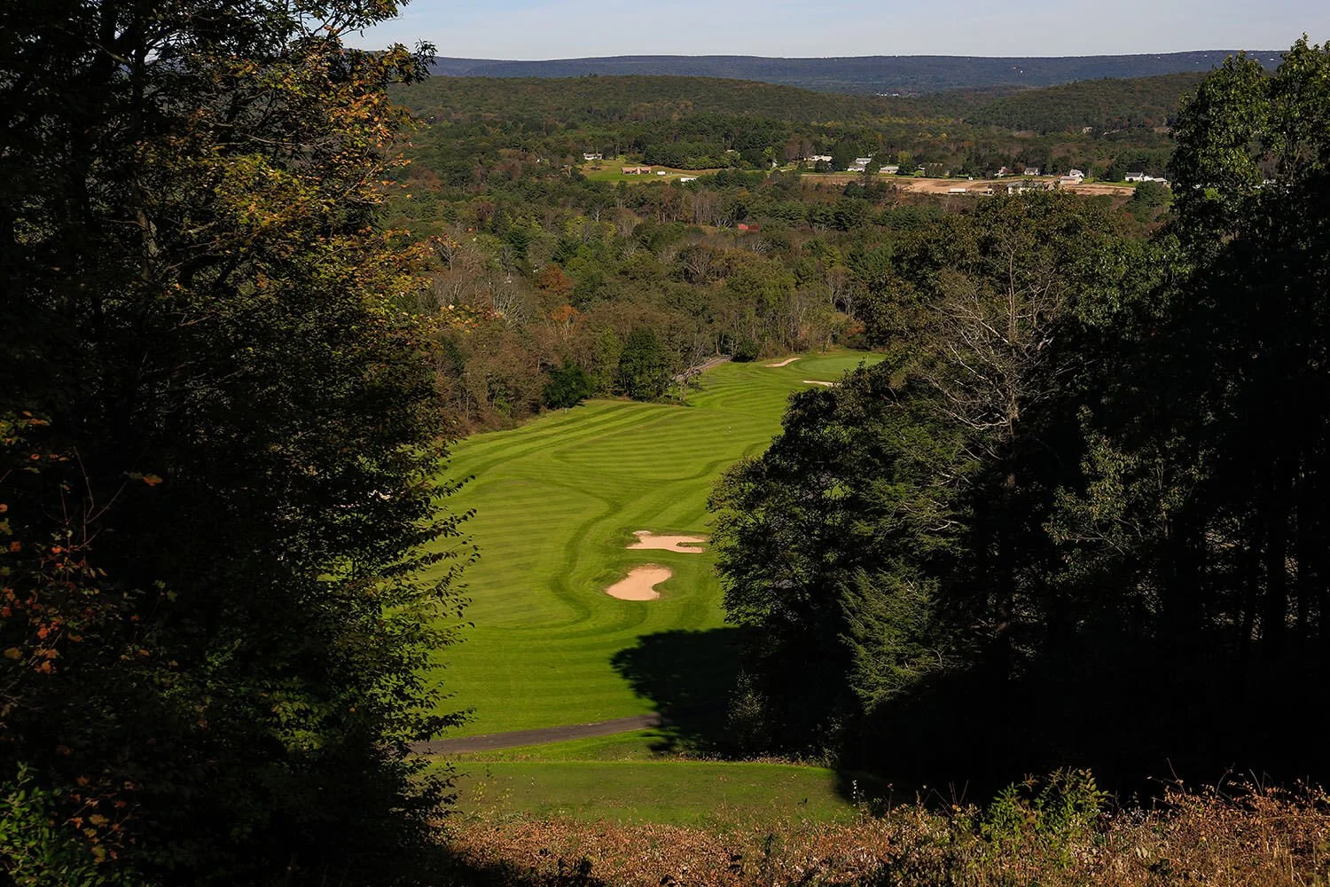 A scenic view of a golf course fairway bordered by trees, with bunkers and a lush green landscape, in a hilly area under a clear sky.