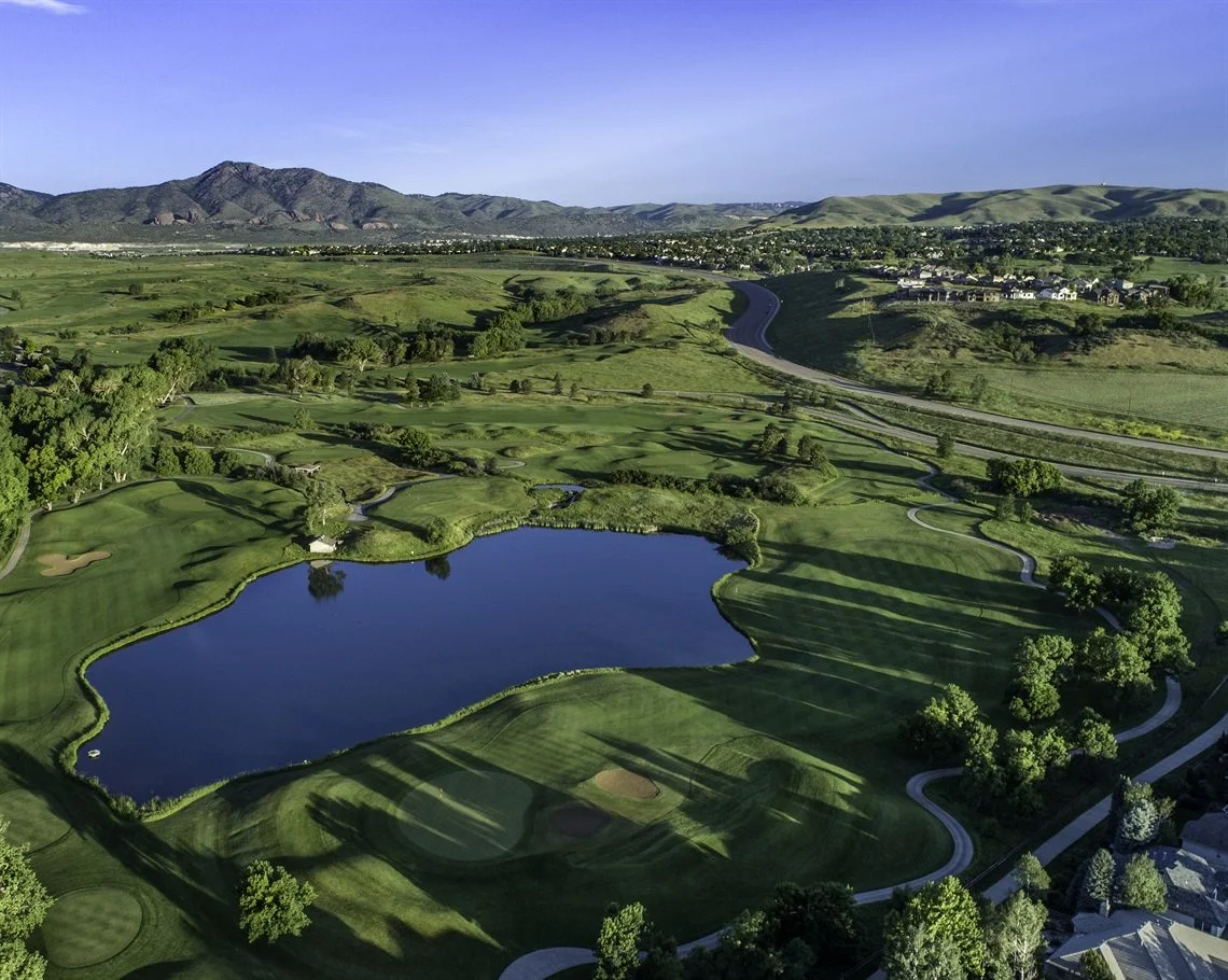 Aerial view of a golf course with a large water hazard, surrounded by lush green fairways and trees, with rolling hills and mountains in the background under a clear blue sky.