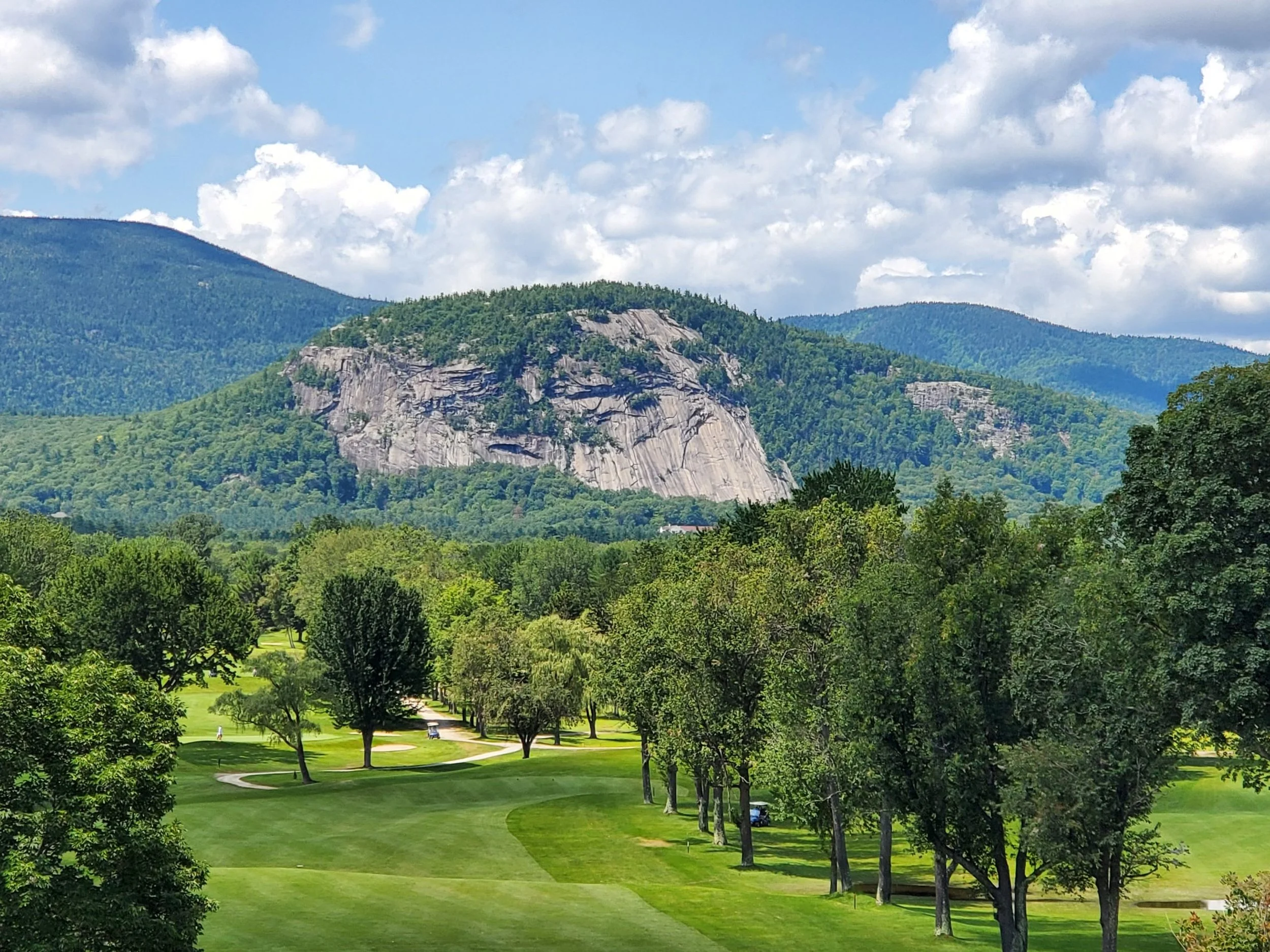 A scenic view of a golf course with green grass and trees, with rocky mountain and hills in the background under a partly cloudy sky.
