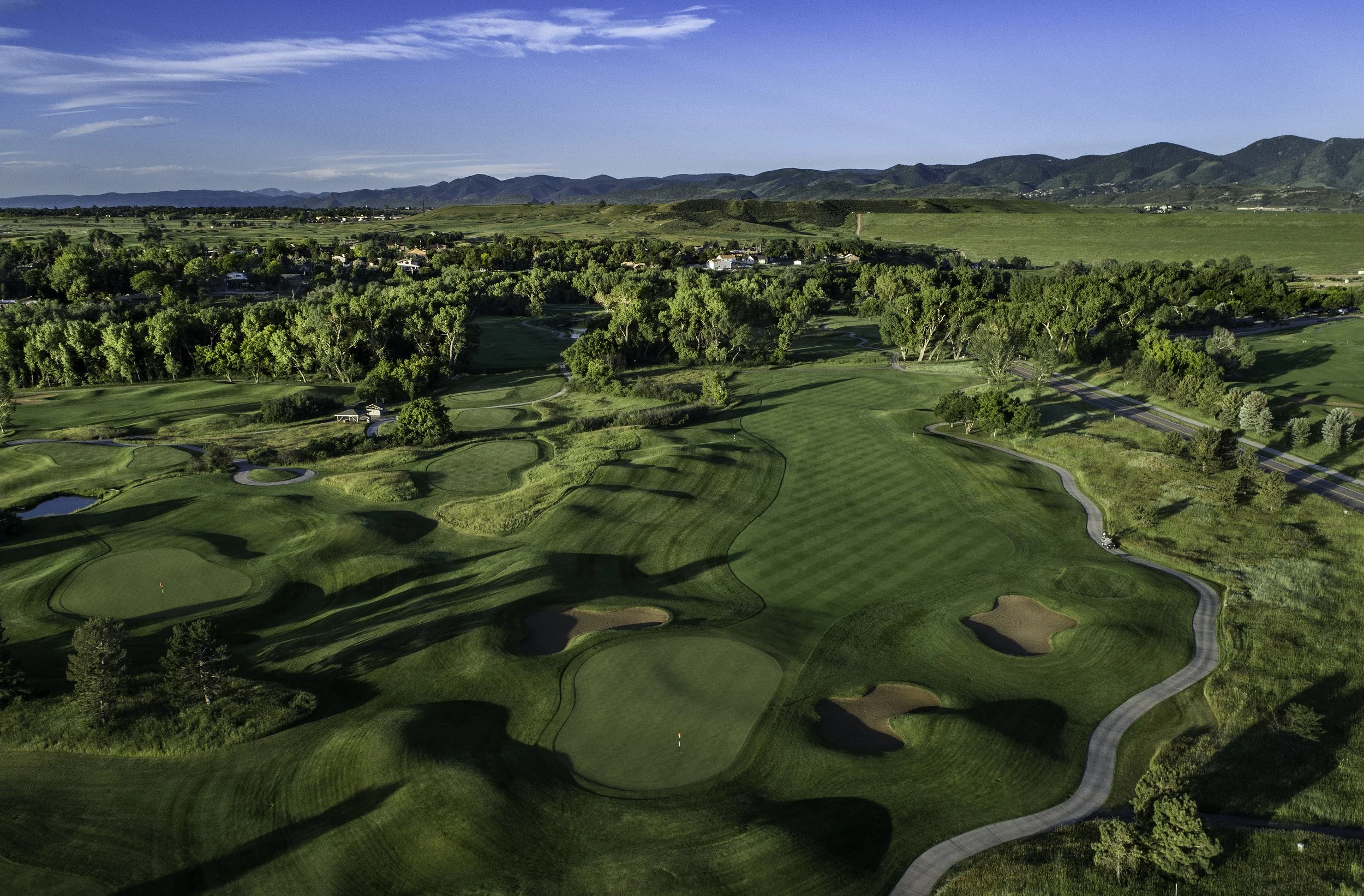 Aerial view of a golf course with well-manicured greens, sand traps, and surrounding trees, with hills and mountains in the background under a blue sky.