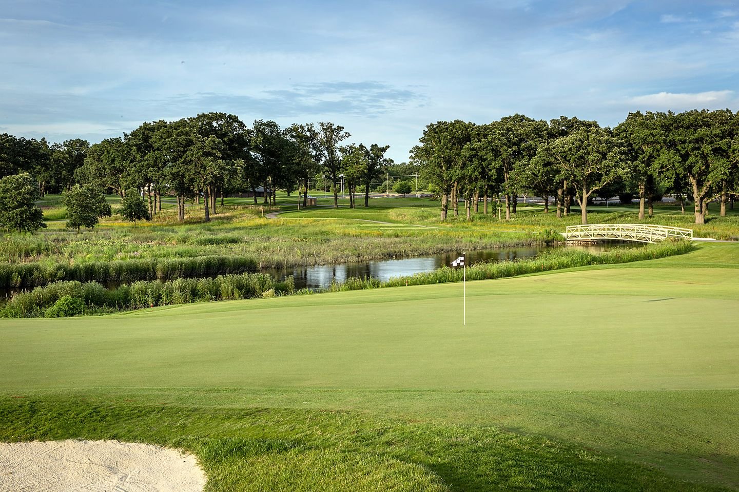 A scenic golf course with a putting green, flag, small pond, trees, and a small white bridge.
