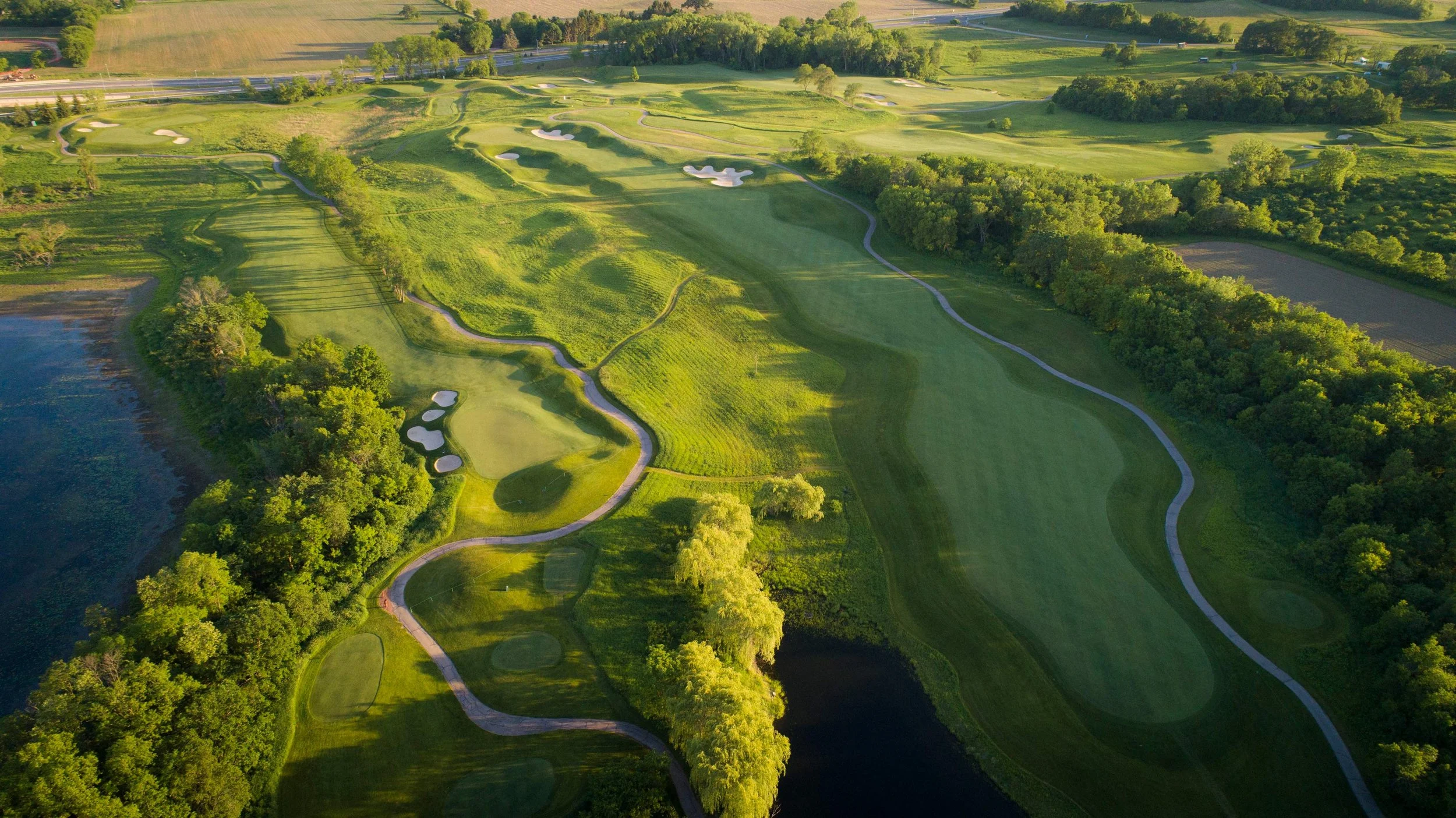 Aerial view of a golf course with multiple fairways, greens, sand traps, water hazards, and surrounding trees, located next to fields and a body of water.