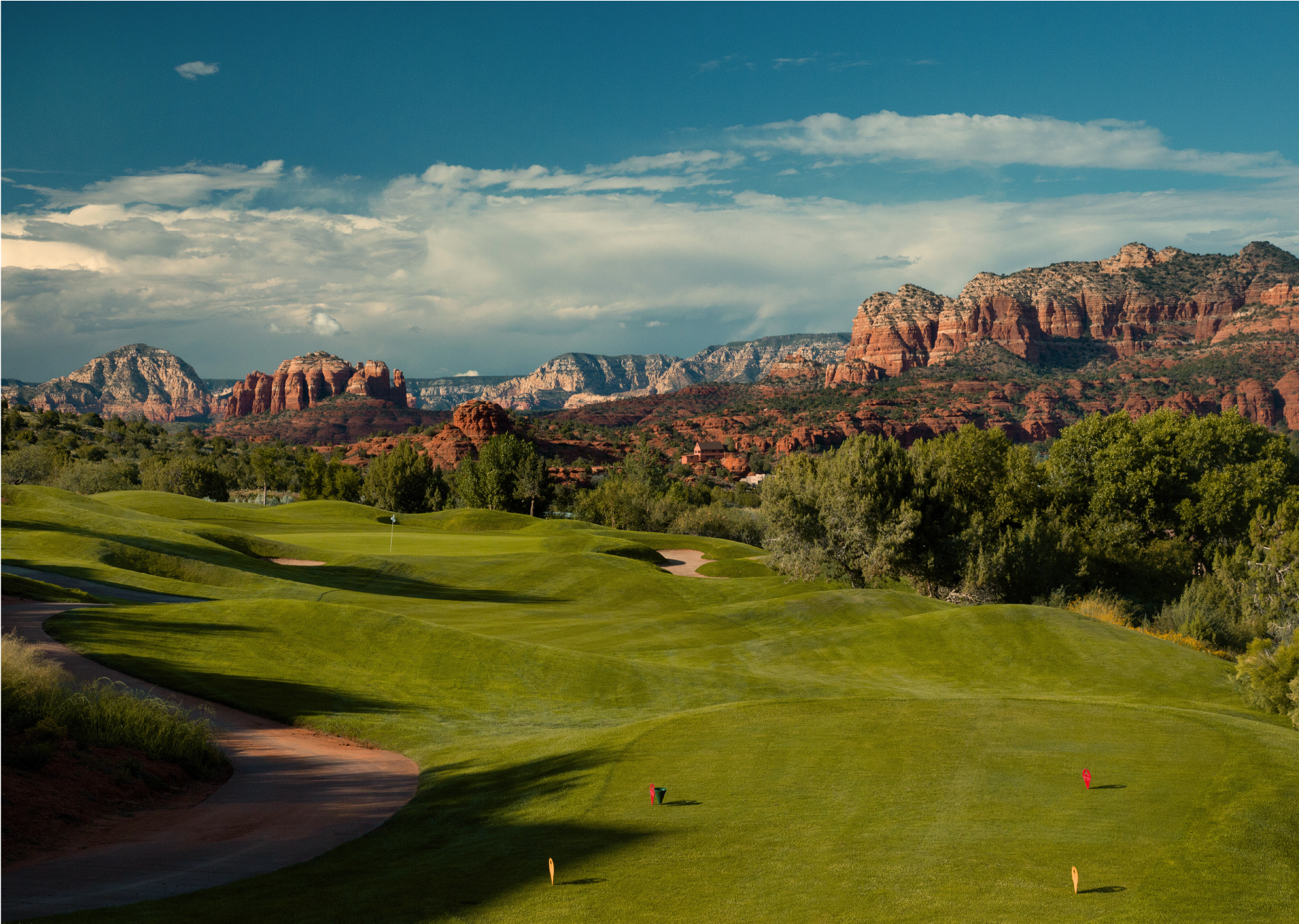 Scenic view of a golf course with green fairways, surrounded by trees, with red rock mountains and a partly cloudy sky in the background.