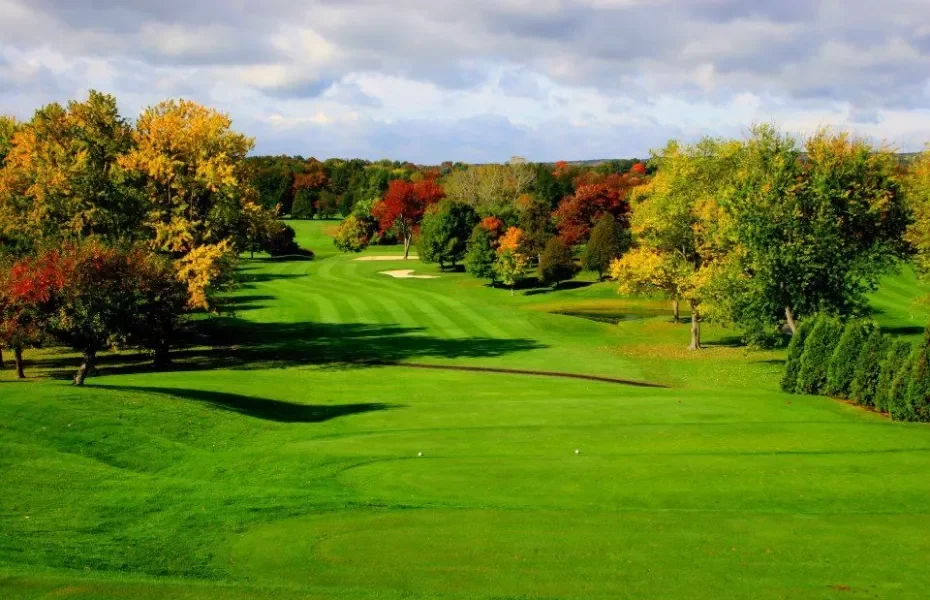 A lush golf course with neatly mowed grass, surrounded by colorful trees in fall foliage, under a partly cloudy sky.