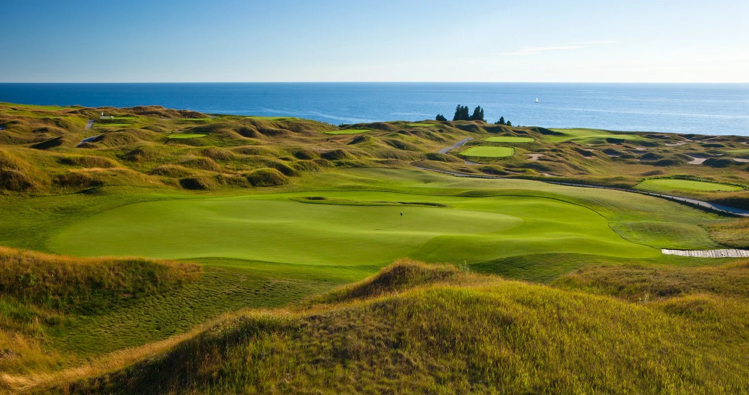 Scenic golf course with green fairways and sand traps near the coastline under a clear blue sky.
