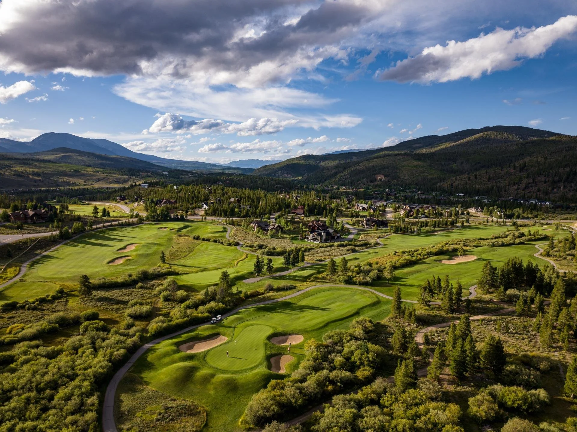 A scenic view of a golf course surrounded by trees, houses, and mountains under a partly cloudy sky.