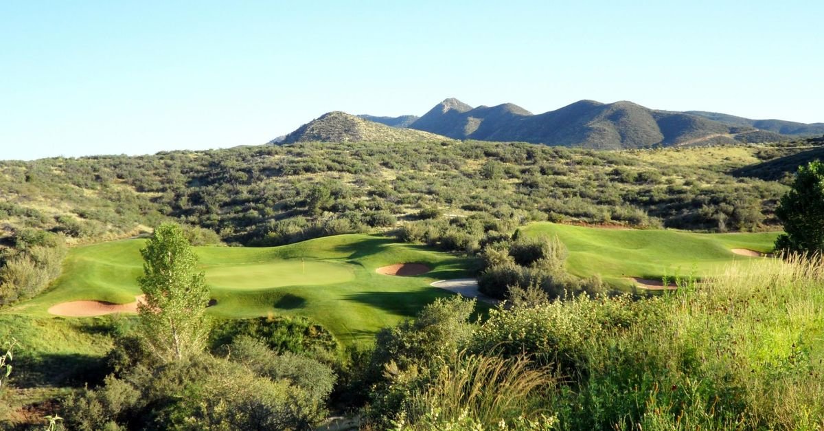 Scenic view of a golf course with green fairways and sand traps, surrounded by bushes and trees, with mountains in the background under a clear blue sky.