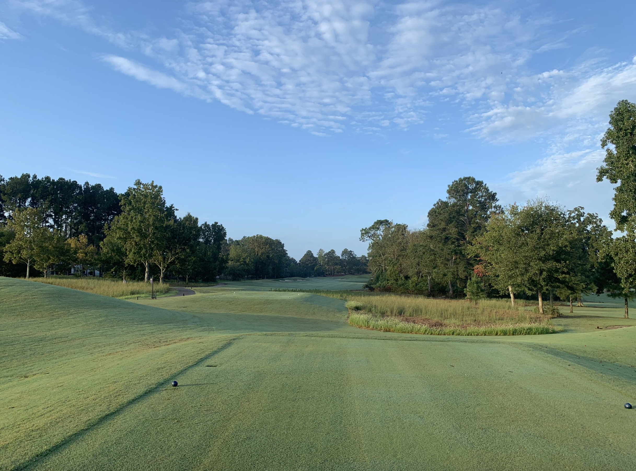A golf course fairway with trees on both sides under a blue sky with scattered clouds.