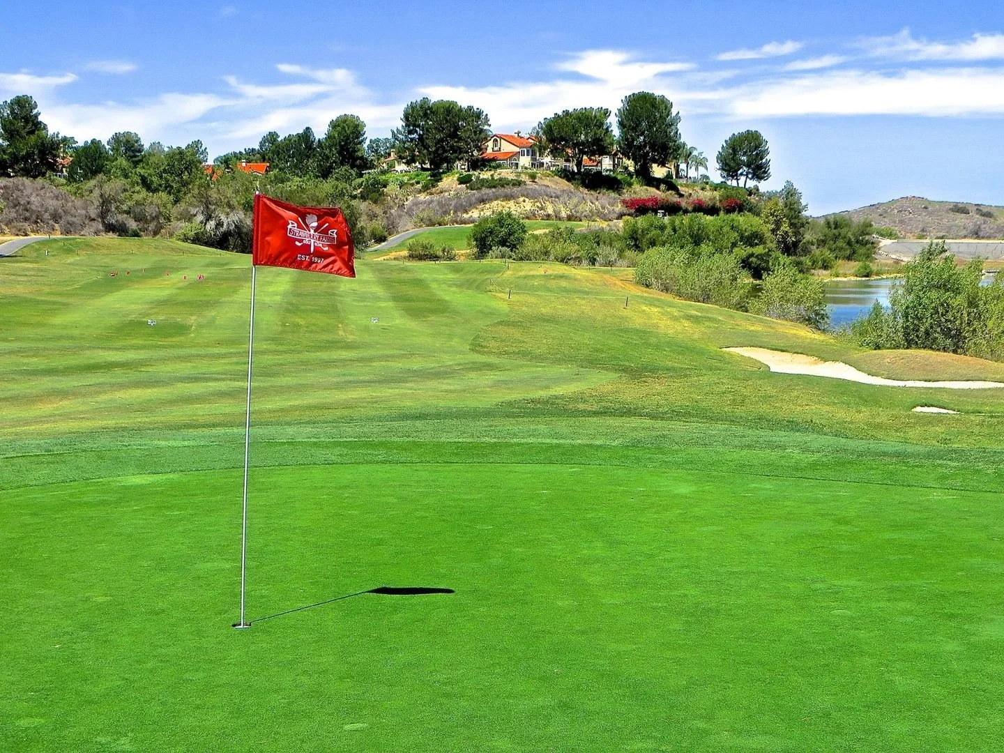 A golf course with a red flag marking the hole, green grass, sand bunkers, water hazards, trees, and houses on a hillside under a blue sky with clouds.